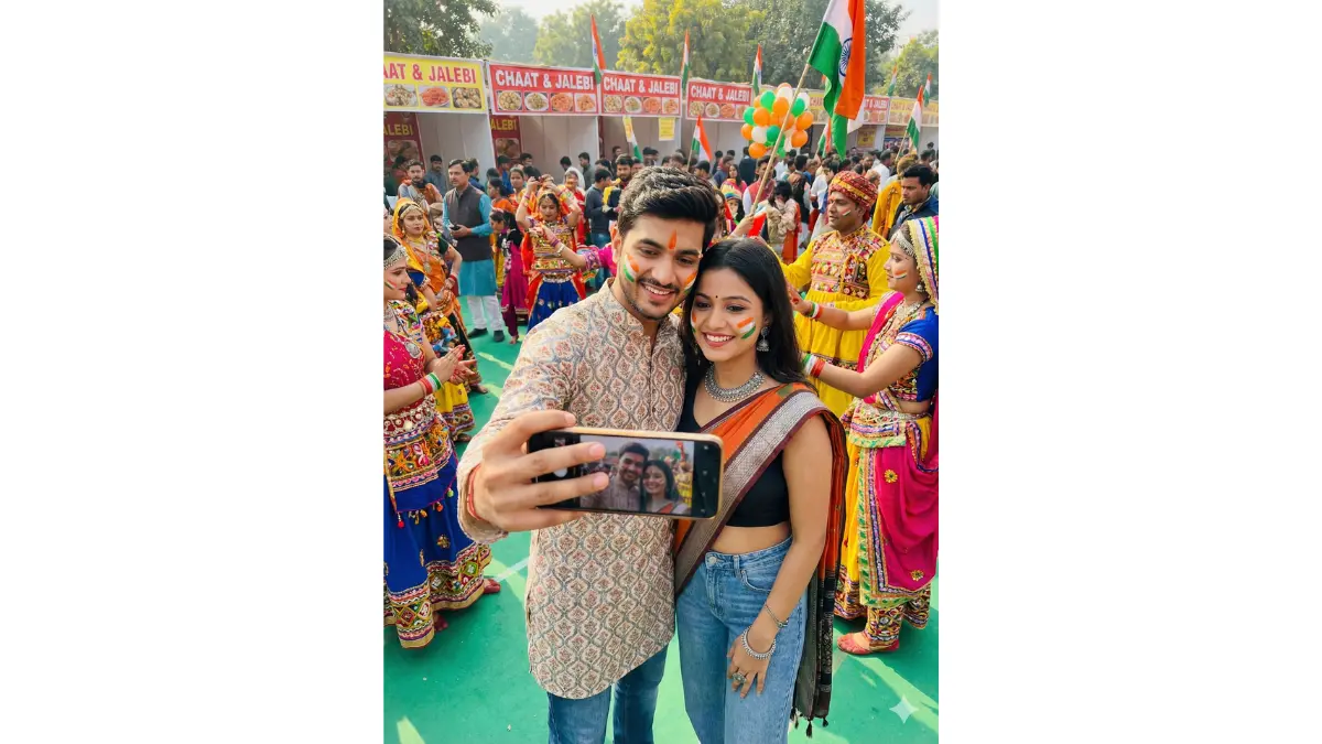 A vibrant modern couple taking a joyful selfie with a smartphone at a bustling Republic Day cultural festival, their faces adorned with artistic tricolor patterns in saffron, white, and green face paint; wearing stylish ethnic-modern fusion: the man in a kurta with contemporary prints over jeans and sneakers; the woman in a modern saree blouse with jeans and ethnic jewelry; surrounded by traditional dancers in colorful costumes performing folk dances, food stalls offering street eats like chaat and jalebi, and a diverse crowd with balloons and flags, highlighting cultural vibrancy and youthful energy.