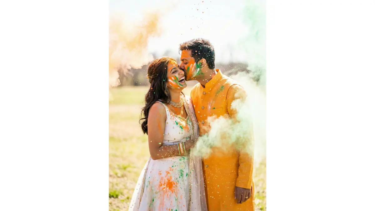 Playful vibrant Republic Day shot, TIGHT MEDIUM-CLOSE SHOT (upper-body, faces hyper-detailed with laughing delight). Open field "Holi of colors" fusion with saffron-white-green powders thrown mid-air. Couple covered in tricolor dust sharing a powder-smeared kiss, her in white lehenga with color bursts, him in saffron bandhgala. High-energy powder clouds, bright daylight bokeh