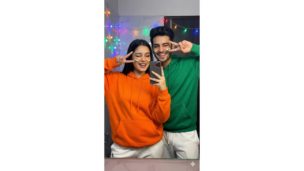 portrait shot of a stylish GenZ couple taking a mirror selfie in matching tricolor outfits – saffron oversized hoodie for her, green for him, white joggers, both making peace signs with small Indian flags painted on their cheeks, playful winks and big grins, bathroom mirror with Republic Day lights reflection, neon glow on faces, viral selfie aesthetic, sharp focus on expressions, makeup details, flag paint, and hoodie textures