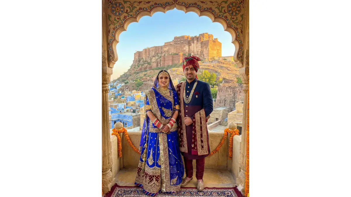 a majestic Jodhpur desert wedding scene while keeping our faces recognizable. Dress the bride in a Rajasthani Poshak-style lehenga or heavy bridal lehenga in royal blue, maroon or pink with traditional Rajasthani jewelry including aad (choker) and rakhdi. Dress the groom in an embroidered sherwani with a Jodhpuri bandhgala jacket and traditional mojari juttis. Replace the background with Mehrangarh Fort's imposing structure, blue city glimpses, and Thar desert landscape. Add dramatic lighting highlighting the fort's architecture. Include royal elements like traditional jharokhas and Rajasthani folk art patterns.