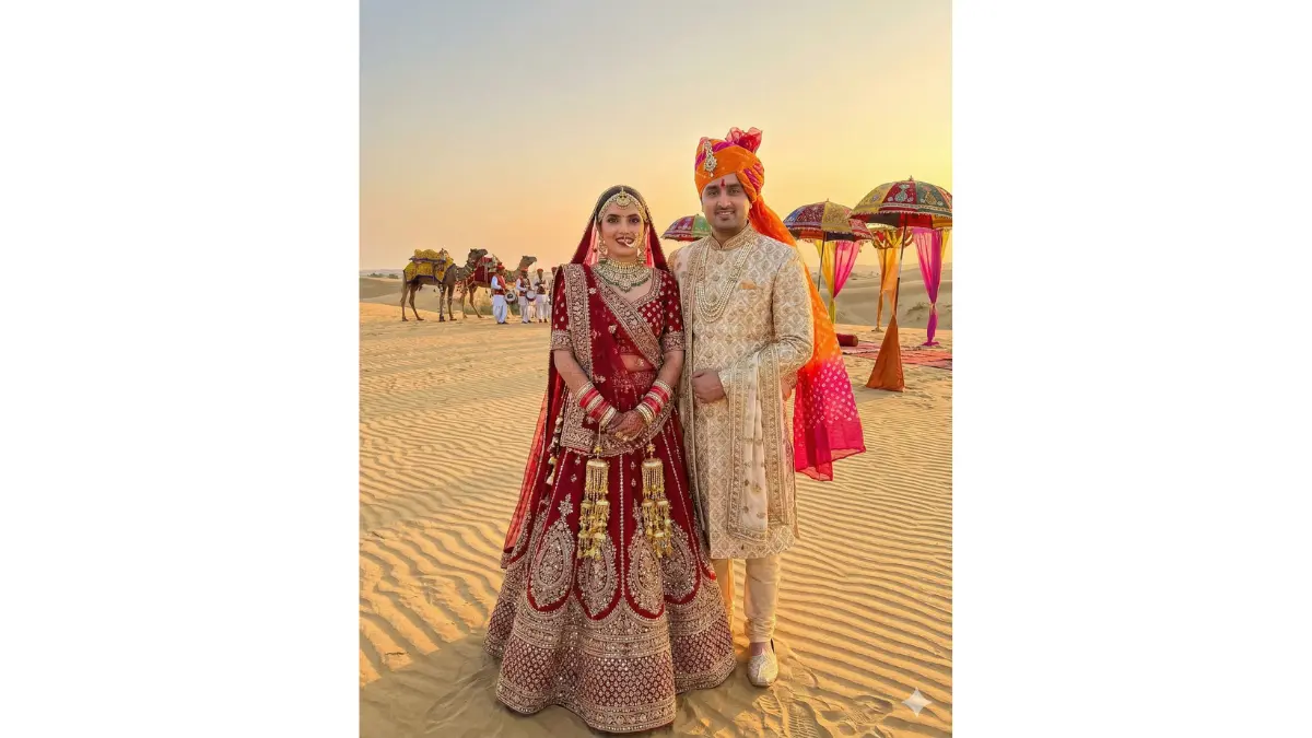 a traditional Rajasthan Thar desert wedding portrait while preserving our exact faces. Dress the bride in a heavily embellished red or maroon bridal lehenga with traditional solah shringar (16 adornments) including kalire, chooda, maang tikka, and jhumkas. Dress the groom in a cream or gold sherwani with a bright Rajasthani pagdi (turban) and traditional kalghi. Change the background to expansive golden sand dunes with a decorated camel caravan in the distance. Add traditional colorful Rajasthani umbrellas and fabric decorations. Include warm sunset lighting creating dramatic shadows on dunes.