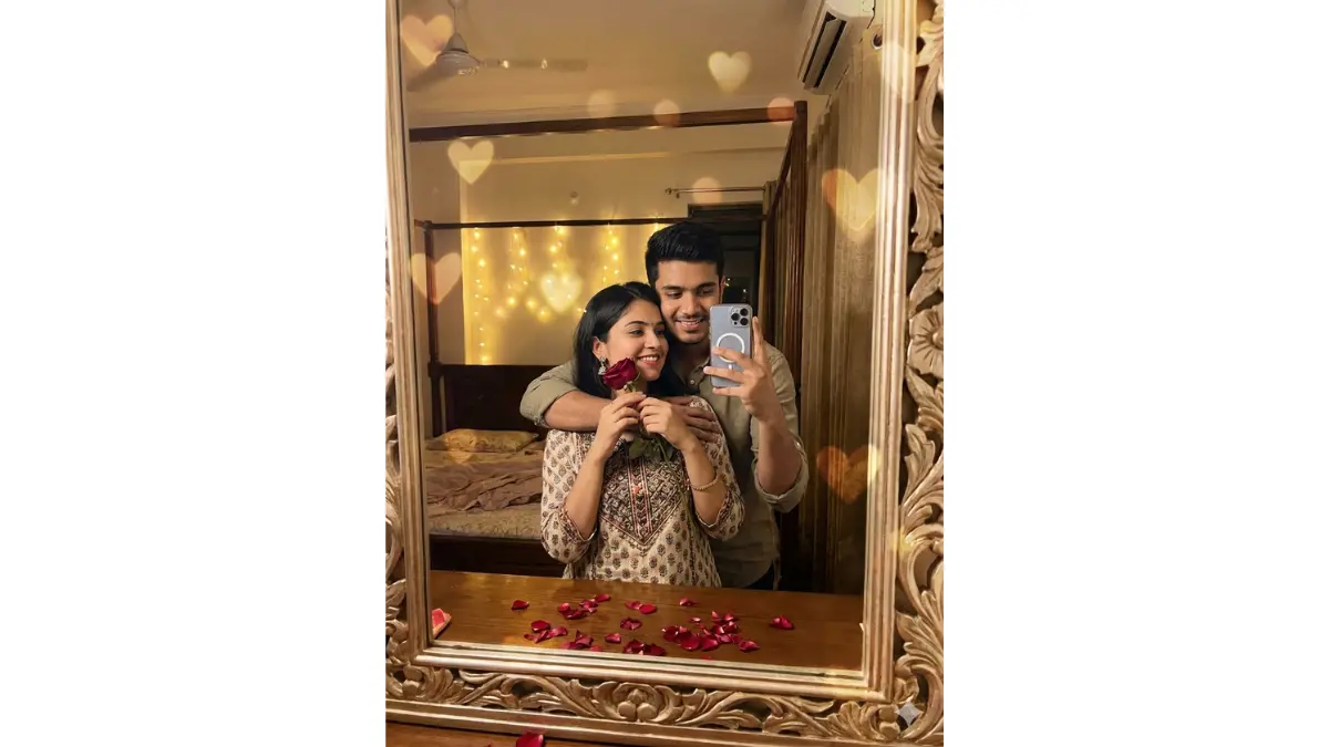a cute Rose Day mirror moment: they take a mirror selfie in a bedroom, girlfriend holding a red rose near her cheek while boyfriend hugs her from behind, both smiling at reflection, preserve mirror composition, exact faces, expressions, outfits, hair, and selfie pose with no alterations, add soft bedroom lighting, scattered petals around mirror, heart bokeh subtly